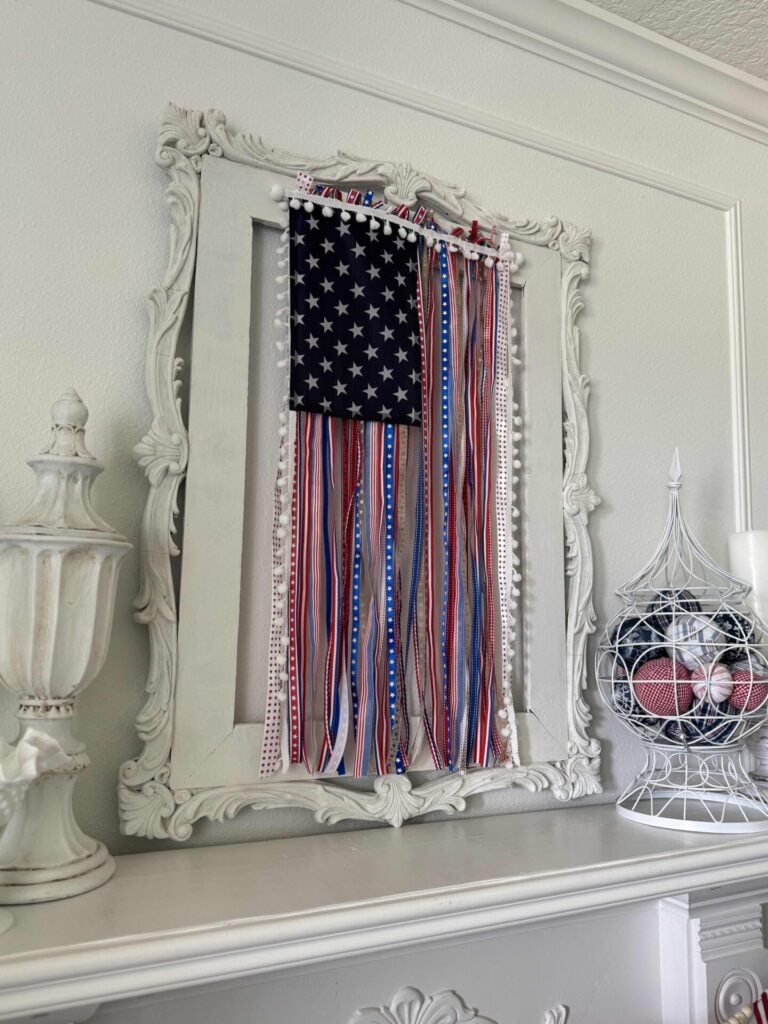 A decorative white-framed display showcases a DIY Patriotic Ribbon Flag. It's hanging over a mantel with white finials and a wire basket filled with ribbon orbs.