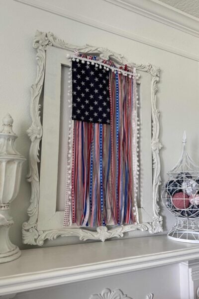 A decorative white frame displays a DIY Patriotic Ribbon Flag, featuring red, white, and blue ribbons with a navy blue section of white stars. The abstract flag design sits on a white mantle adorned with ornate decor.