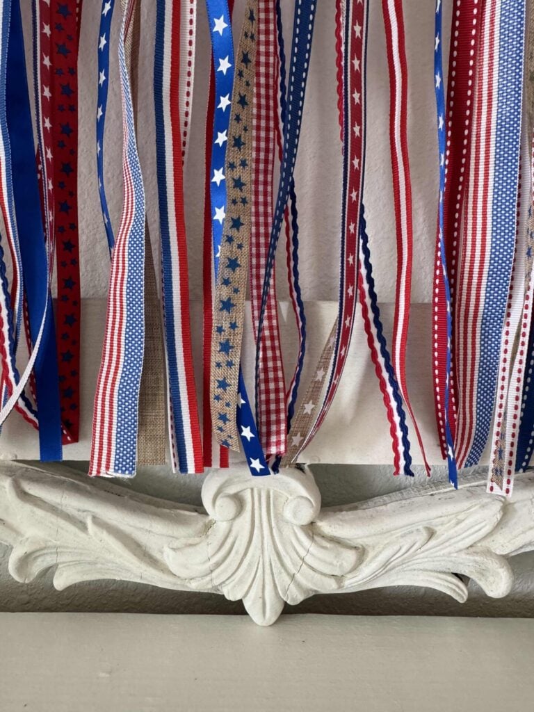 Red, white, and blue ribbons with stars, stripes, and checks hang in a row against a decorative white carved surface, perfect for crafting a DIY Patriotic Ribbon Flag that captures the spirit of Americana.
