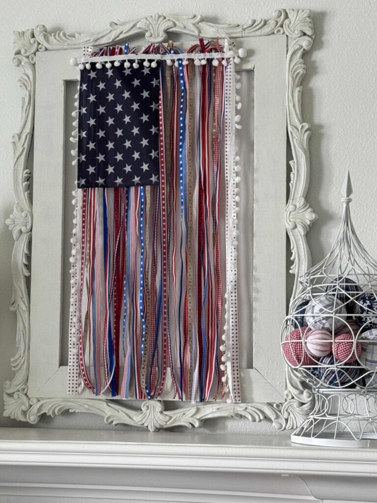 A decorative frame showcases a DIY Patriotic Ribbon Flag, with long red, white, and blue ribbons hanging down. Nearby, a white wire birdcage displays matching fabric orbs, all set on a bright white mantel.