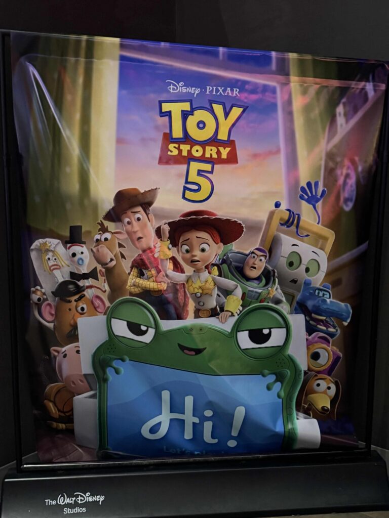 A poster for “Toy Story 5” shows Woody, Jessie, Buzz Lightyear, and other toy characters together, sitting behind a green toy frog holding a blue sign that says Hi! The Disney Pixar logo is at the top.