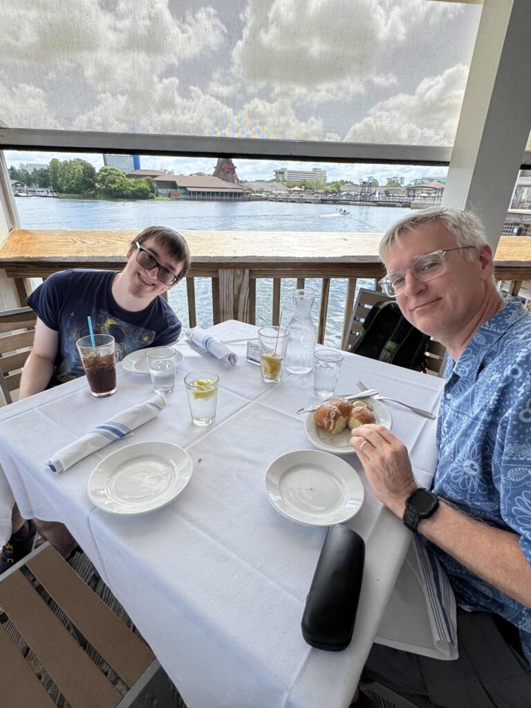 Two people sit at a riverside restaurant table with drinks and plates, one eating a sandwich. Large windows show a river, trees, and buildings under a cloudy sky in the background.