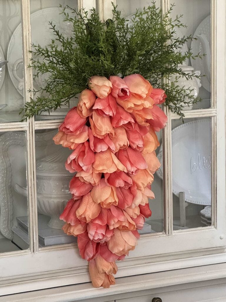 A decorative arrangement of pink and peach tulips shaped like a carrot, topped with green foliage, hangs on a white cabinet with glass doors and displayed dishes inside.