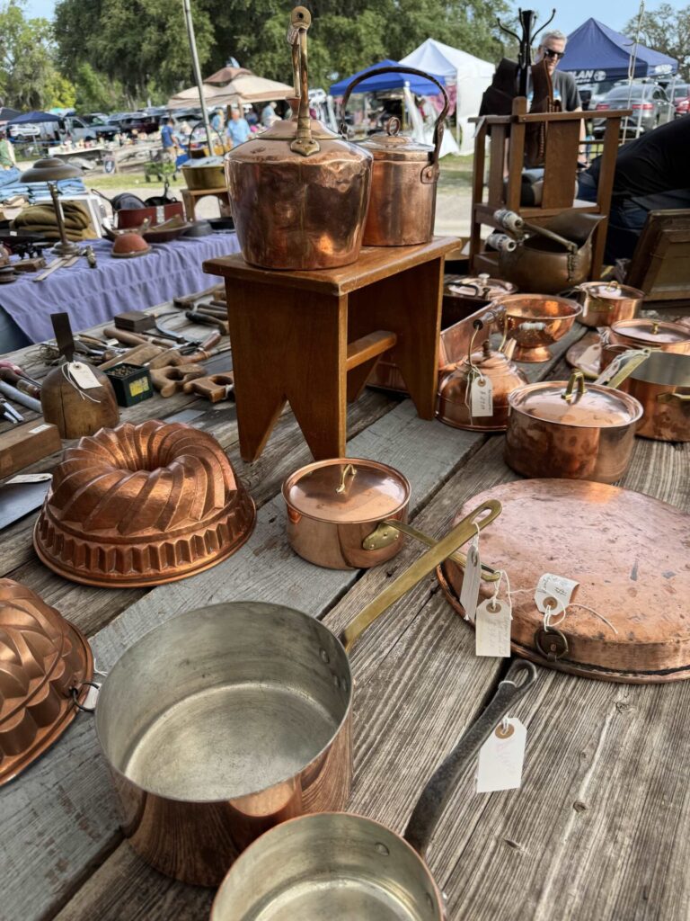 A variety of vintage copper pots, pans, and kitchenware with price tags are displayed on a wooden table at an outdoor market. Other vendor booths and tents are visible in the background.