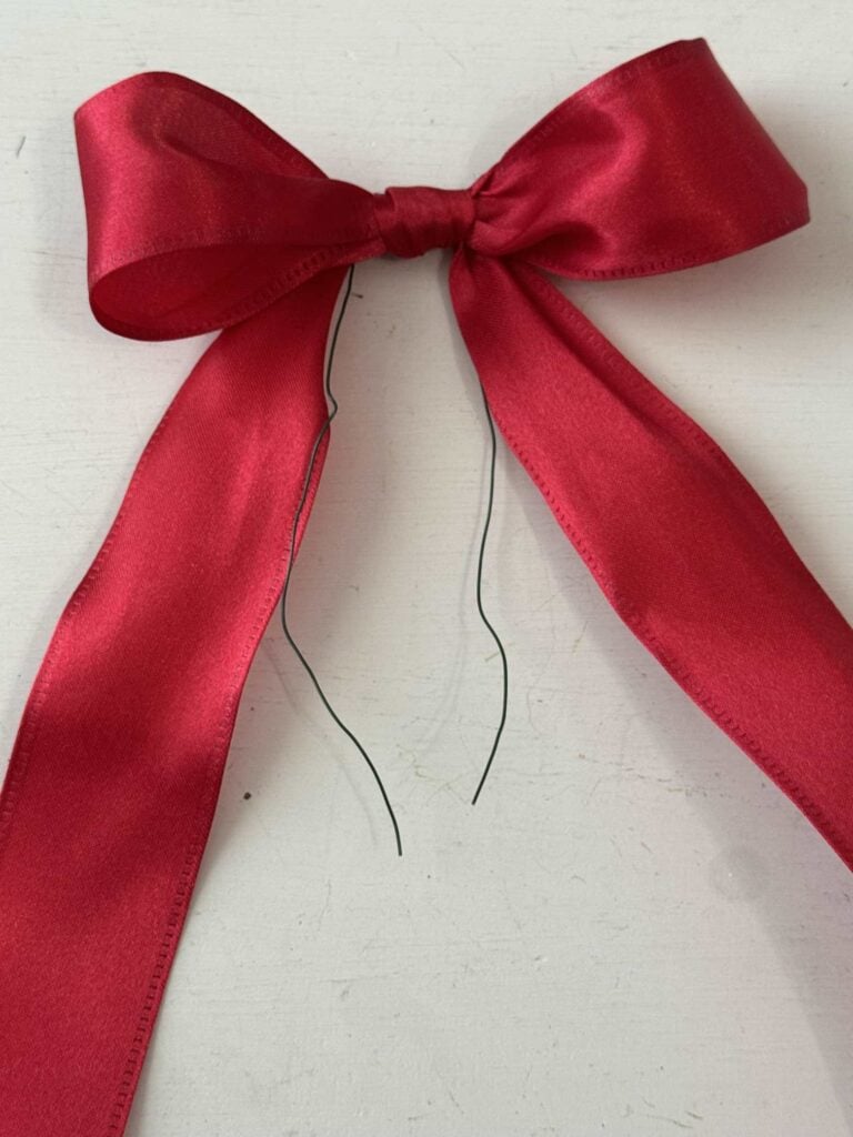 A red satin ribbon tied into a bow with two long tails, lying on a white surface. Thin black wires hang down from the center of the bow.