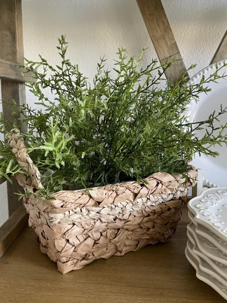 A woven basket filled with green faux plants sits on a wooden shelf next to DIY Fabric Orbs and a stack of white decorative plates with embossed patterns.