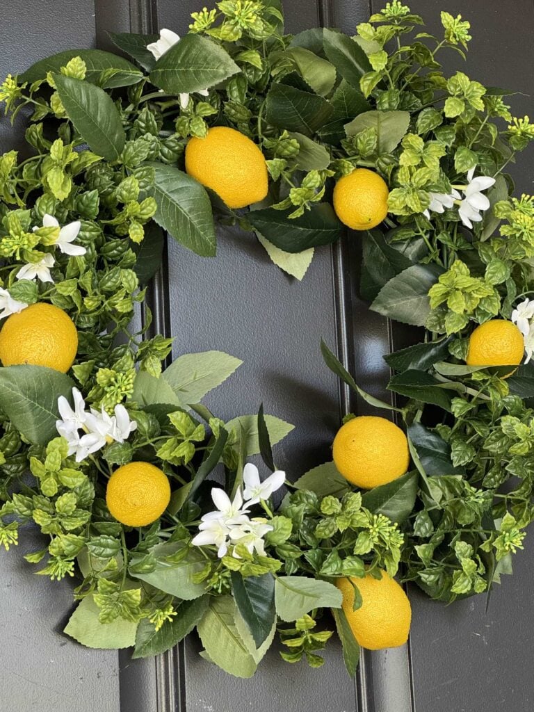A decorative DIY Frame Wreath made of green leaves, small white flowers, and several bright yellow artificial lemons, hanging on a dark-colored door.