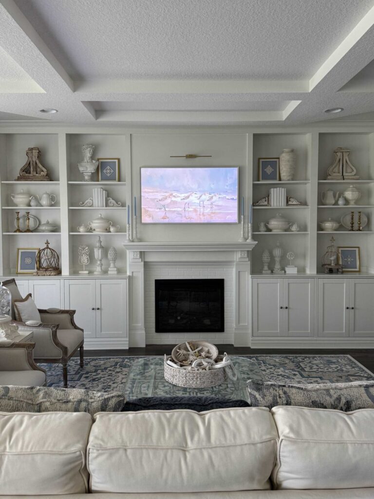 A stylish living room with a white sofa, glass coffee table, fireplace, and built-in shelves displaying decor. Above the mantle, a TV displays landscape art. Neutral tones and blue accents create a serene, elegant atmosphere.