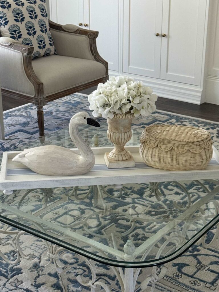 A glass coffee table with a white tray displaying a wooden swan, a cream vase with white flowers, and a woven basket. Behind the table is a beige armchair with a blue patterned pillow and a blue patterned rug.