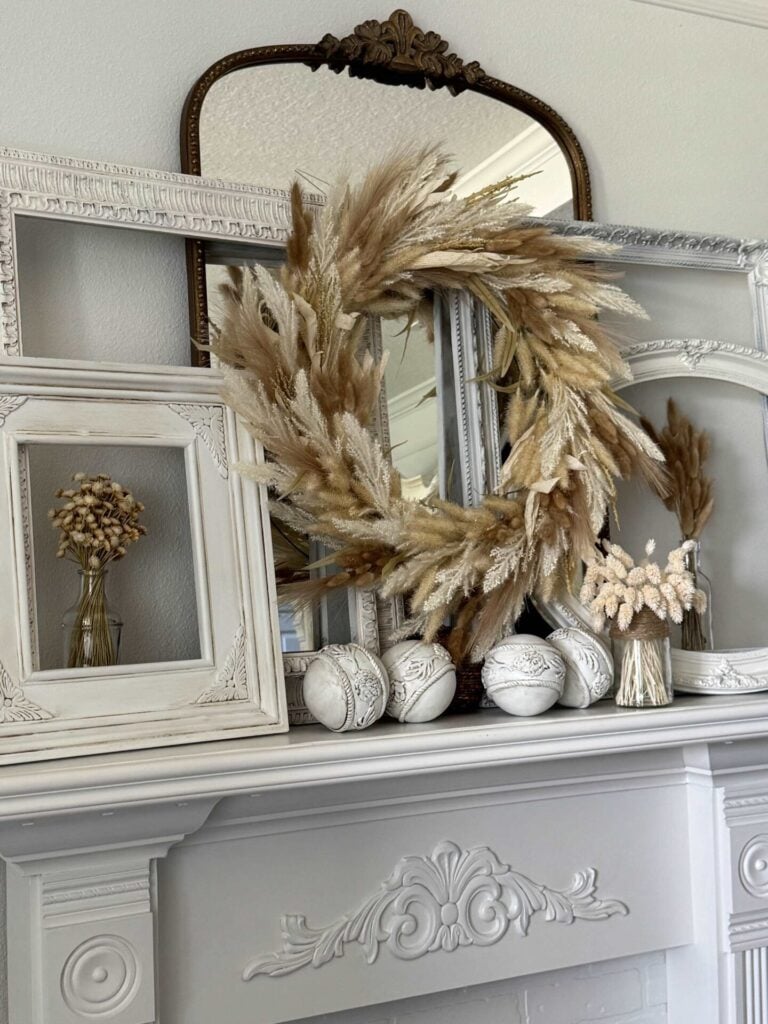 A decorative mantel features empty white frames, a large DIY frame wreath made of dried grass, three white textured orbs, a small vase with dried flowers, and a large antique mirror in the background.