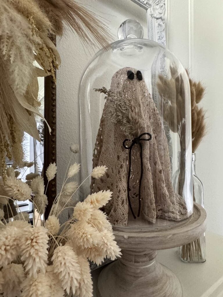 A crochet ghost with black button eyes and a small bouquet stands under a glass dome on a wooden base, surrounded by dried beige grasses, DIY fabric orbs, and neutral-toned decor.
