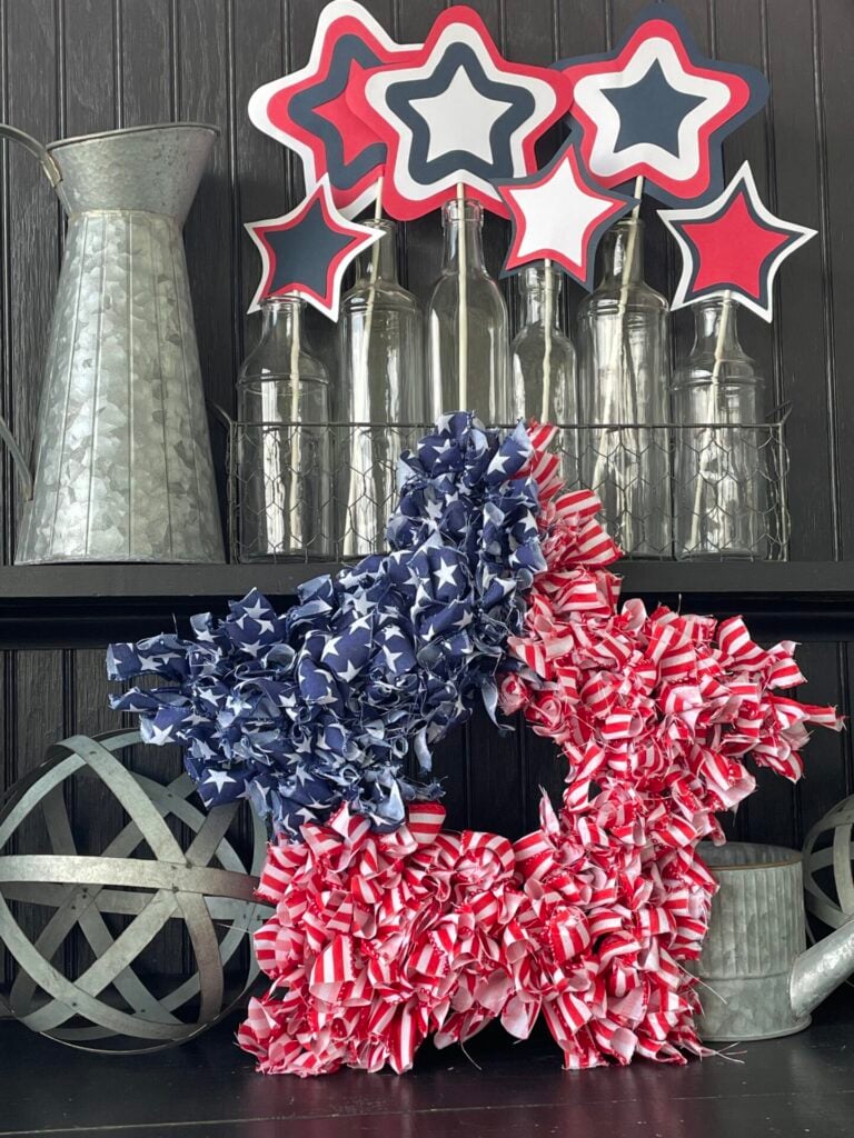 A patriotic star wreath with blue and white stars on one half and red and white stripes on the other sits on a shelf, surrounded by bottles, metal pitchers, a DIY patriotic garland, and red, white, and blue star decorations.