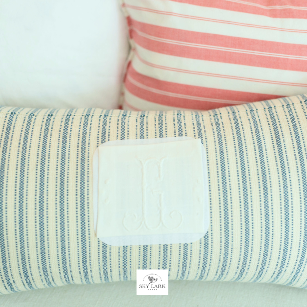 A cylindrical pillow with blue and white stripes and a white patch featuring an embroidered letter G, placed in front of a pink and white striped cushion.