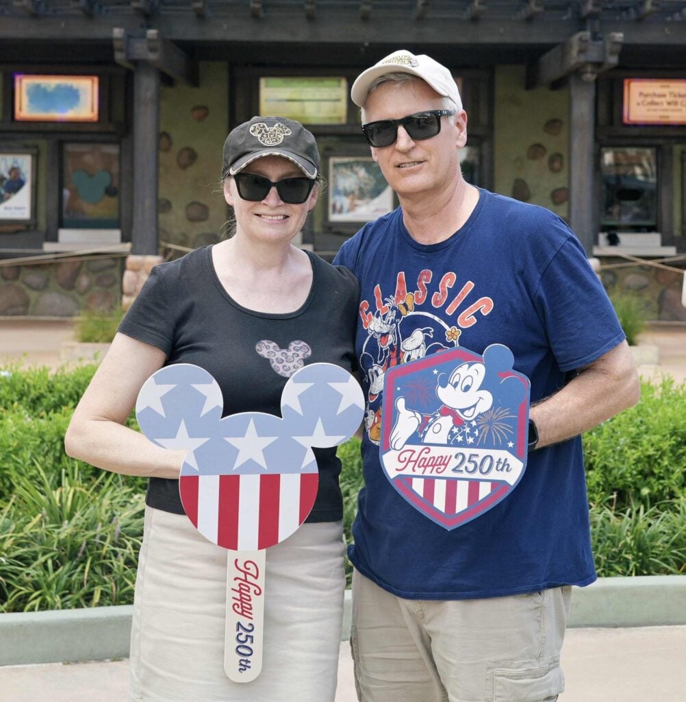 A smiling couple in casual clothes holds Disney-themed signs that say Happy 250th in front of a theme park building with greenery and posters in the background-part for our Seasonal Sentiments July Rewind.
