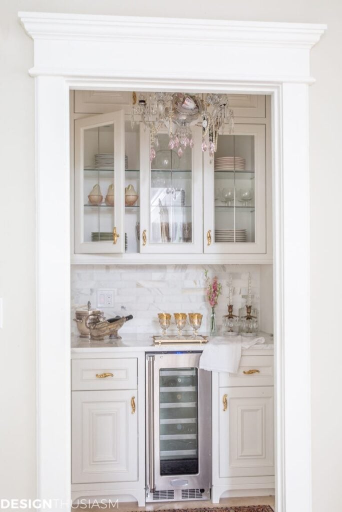 Elegant white butler’s pantry with glass cabinets, gold hardware, marble countertop, wine fridge, and crystal chandelier. Decorative glassware and plates displayed on shelves complete the timeless look.