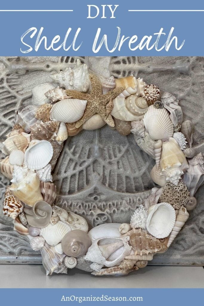 A handmade DIY Shell Wreath adorned with seashells and a starfish is displayed against a textured gray background. The image features “DIY Shell Wreath” at the top and “AnOrganizedSeason.com” at the bottom.