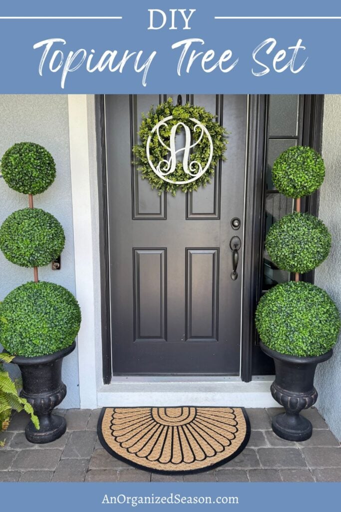 A front door with a wreath featuring a letter A, two stacked DIY topiary trees in black planters on each side, and a semicircle doormat on a stone porch.