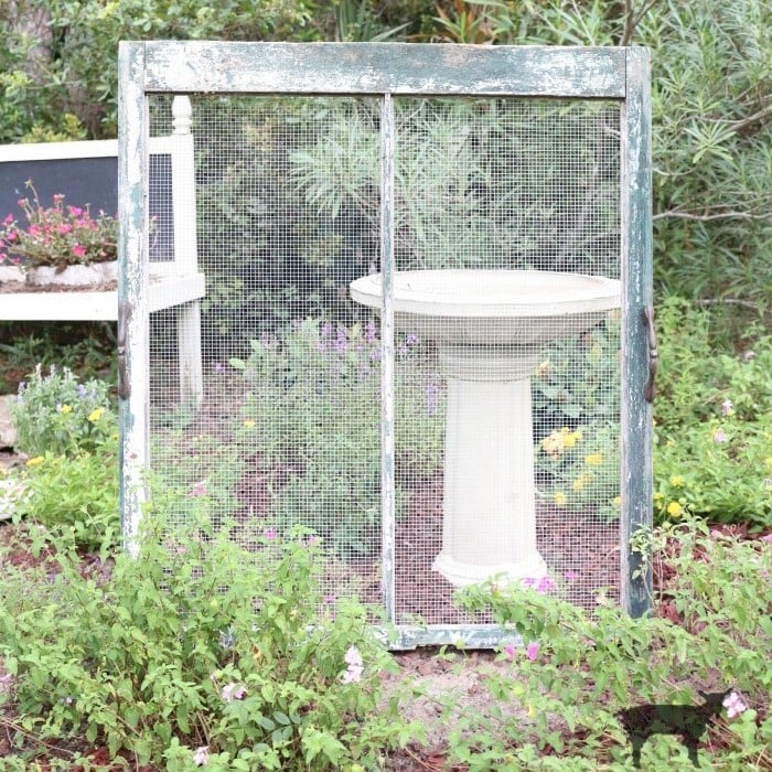 Old window screen frame placed vertically in a garden, with a white birdbath and blooming flowers in the background, creating an illusion of a framed outdoor scene—perfect inspiration for learning how to hang flower pots and add unique charm to your outdoor space.