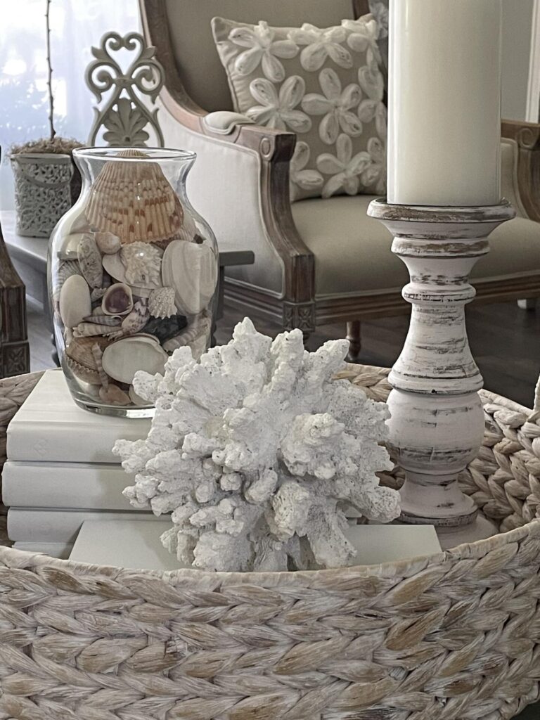 A coastal-themed living room scene beautifully showcases summer decor ideas with a wicker tray, stacked white books, a white coral, and a glass vase of seashells. A distressed white candle holder adds charm, while an armchair with a sand dollar pillow in the background completes this breezy seaside-inspired space.