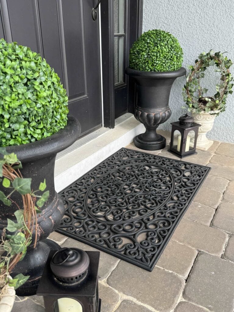 A decorative black doormat on a stone porch, flanked by two potted artificial boxwood plants and a charming DIY topiary tree, alongside two black lanterns with white candles, and a small ivy wreath on a pedestal near a black door.