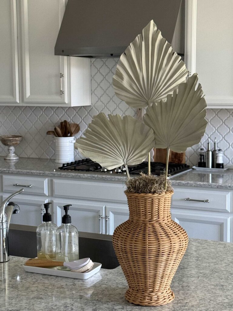 A wicker vase with three dried palm leaves sits on a marble kitchen counter, offering subtle inspiration for Summer Decor Ideas. In the background are white cabinets, a stove, and a backsplash with a geometric tile pattern. Soap dispensers are near the sink.