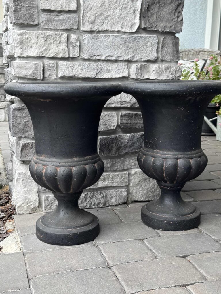 Two black urn-shaped planters with wide rims are placed side by side on a stone patio in front of a gray brick wall, touching at the top edges.