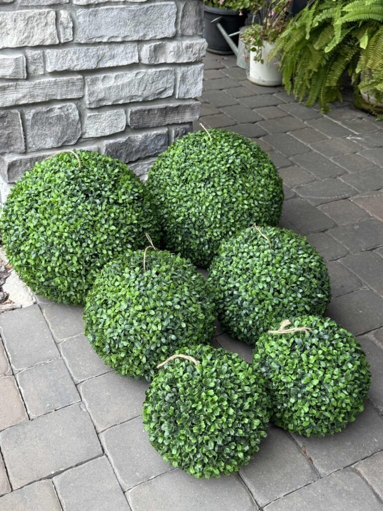 Six artificial boxwood topiary balls of varying sizes are arranged on a stone patio, near a brick wall and some green ferns in white pots.