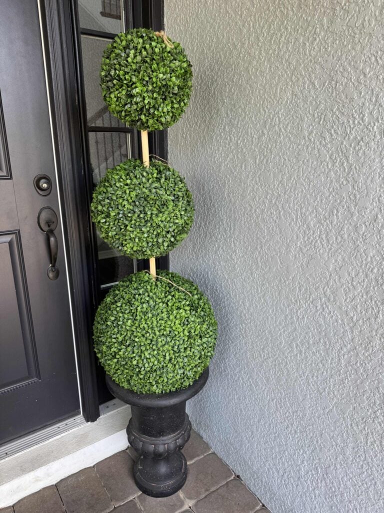 Three stacked artificial green topiary balls in a black planter sit by a gray textured wall and a black door on a brick floor, creating the look of an elegant DIY Topiary Tree.