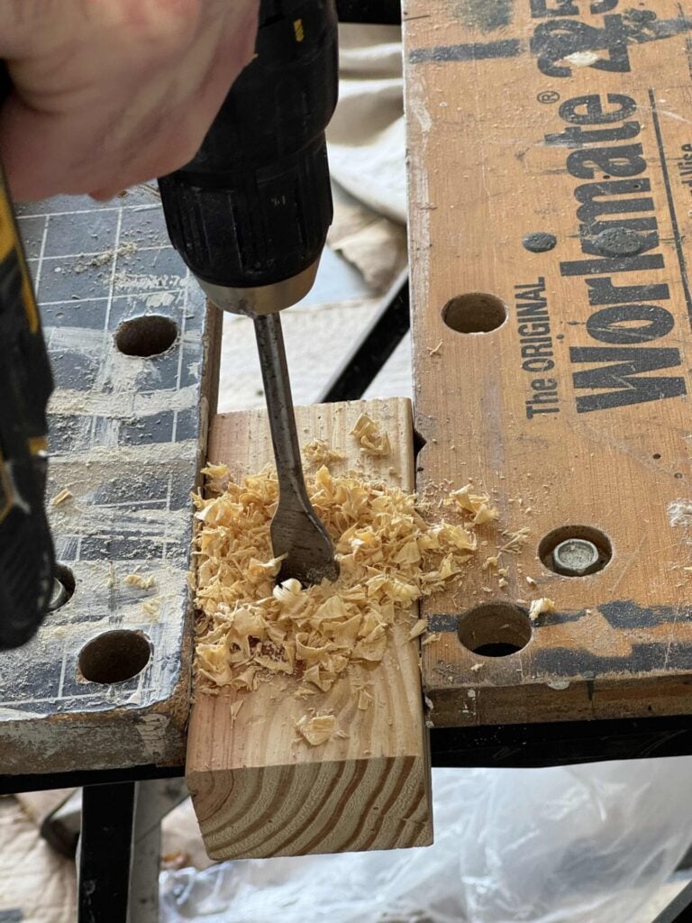 A person uses a power drill with a spade bit to drill into a piece of wood, creating wood shavings on a workbench labeled Workmate.