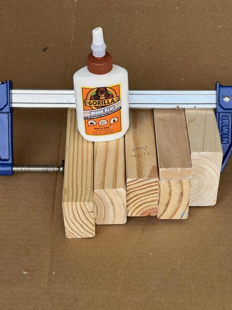 A bottle of Gorilla Wood Glue sits on clamped wooden blocks, held tightly together by a blue Irwin clamp on a brown surface.