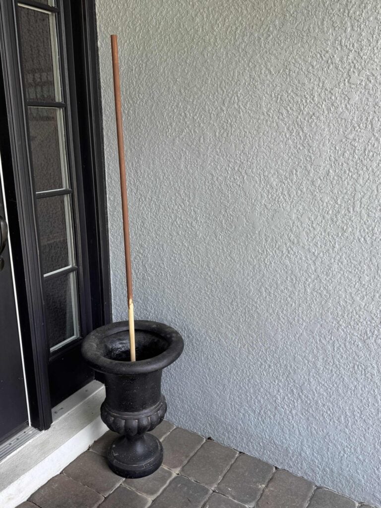 A tall, brown stick is standing upright in an empty, black decorative planter placed in the corner by a textured gray wall and a glass-paneled door on a stone-tiled floor.