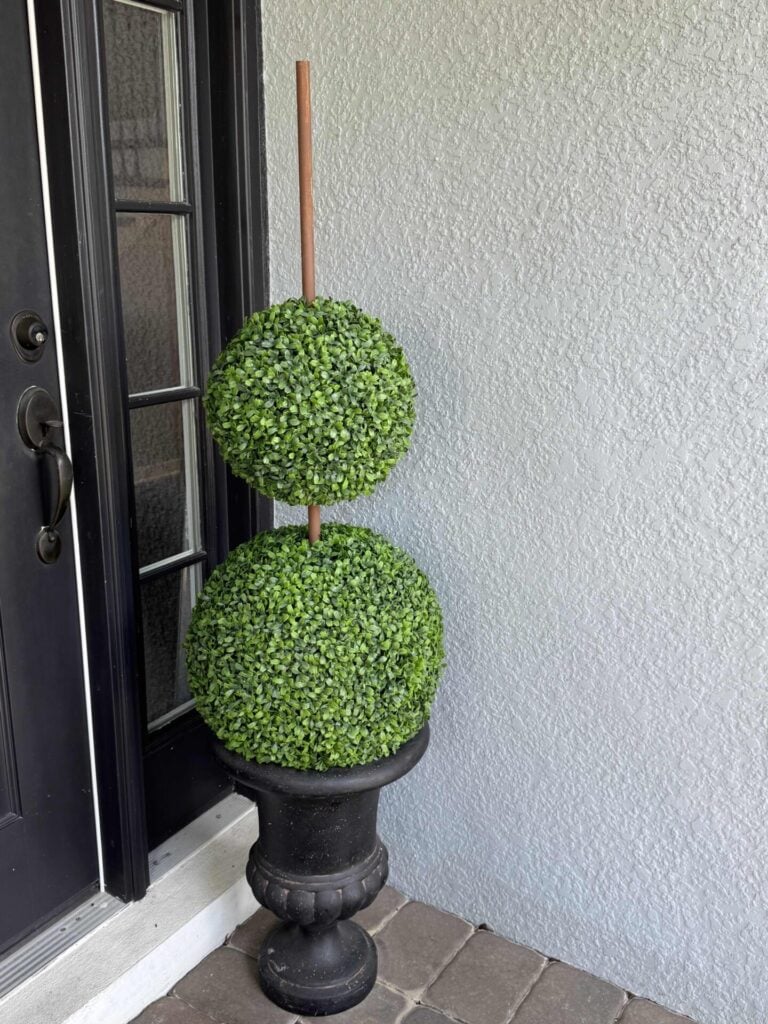 A black planter holds two artificial green topiary balls stacked vertically on a wooden stick, placed next to a white textured wall and a black door on a stone-tiled floor.