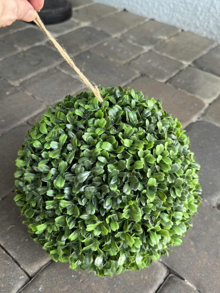 A hand holds a string attached to a round orb made of artificial green leaves, with gray stone tiles in the background.