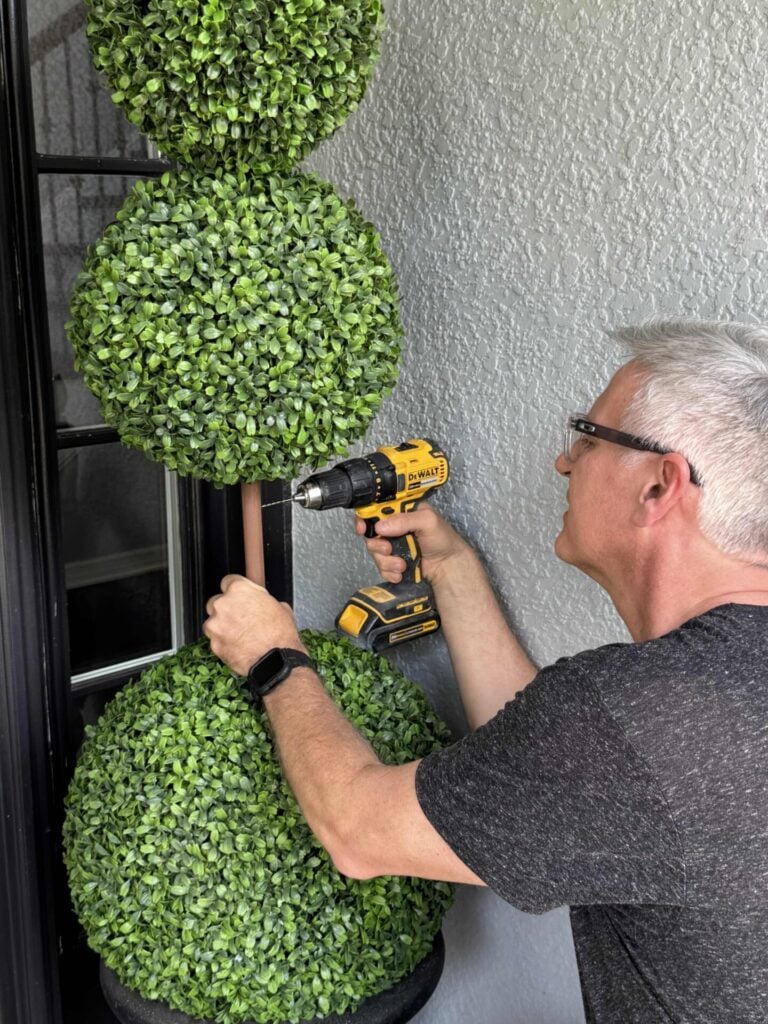 A man uses a yellow power drill to put a hole in the trunk of an artificial plant with three green spheres—against a gray textured wall.