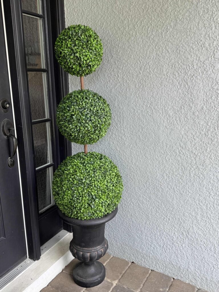 A DIY topiary tree with three stacked green spheres in a black planter stands on a tiled porch next to a black door and a white textured wall.
