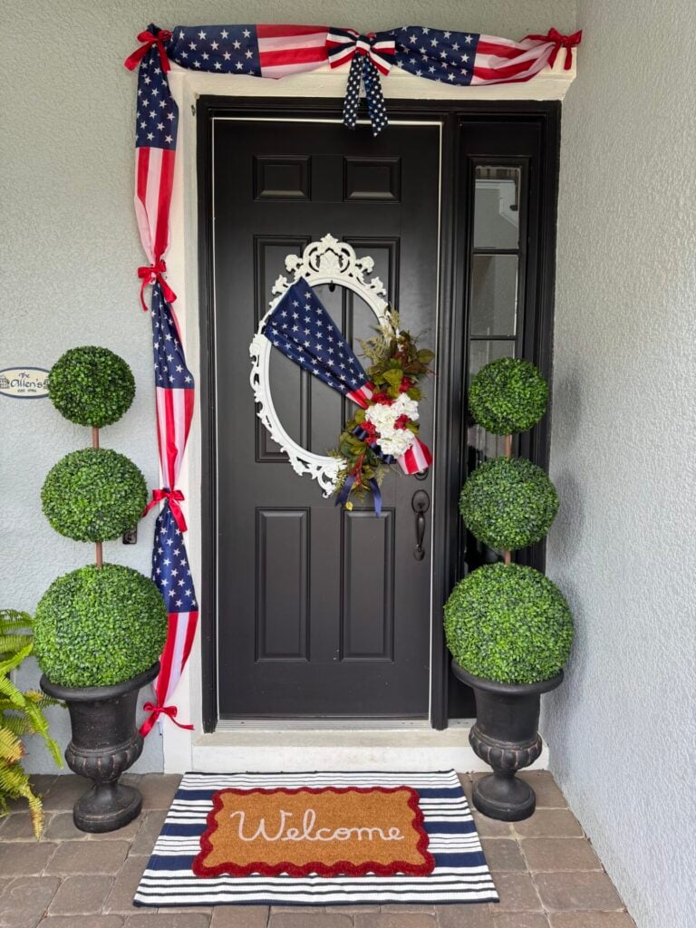 A black front door with patriotic red, white, and blue decorations, including an American flag bow and wreath. Two DIY topiary trees flank the door. A red “Welcome” mat sits on a striped rug at the entrance.