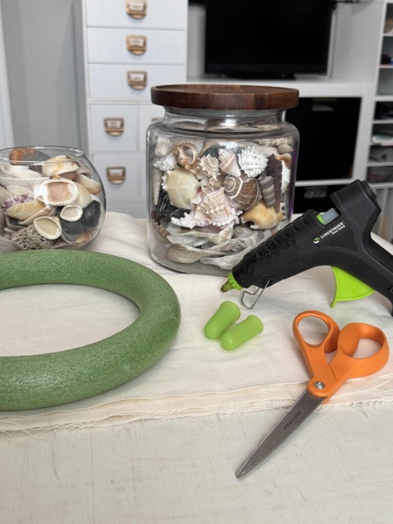 A crafting setup includes a foam wreath, a jar and bowl of seashells, a hot glue gun, green finger protectors, and orange scissors arranged neatly on a table with a light cloth.