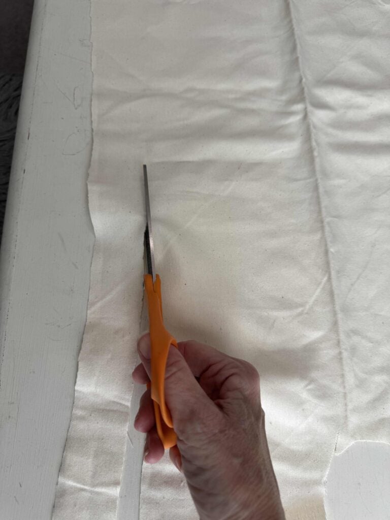 A hand holding orange-handled scissors is cutting a piece of off-white fabric laid out on a white surface.