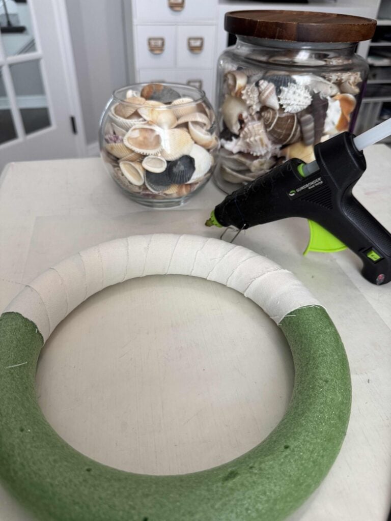 A green foam wreath is partially wrapped in white ribbon and sits on a table near a hot glue gun and jars filled with assorted seashells.