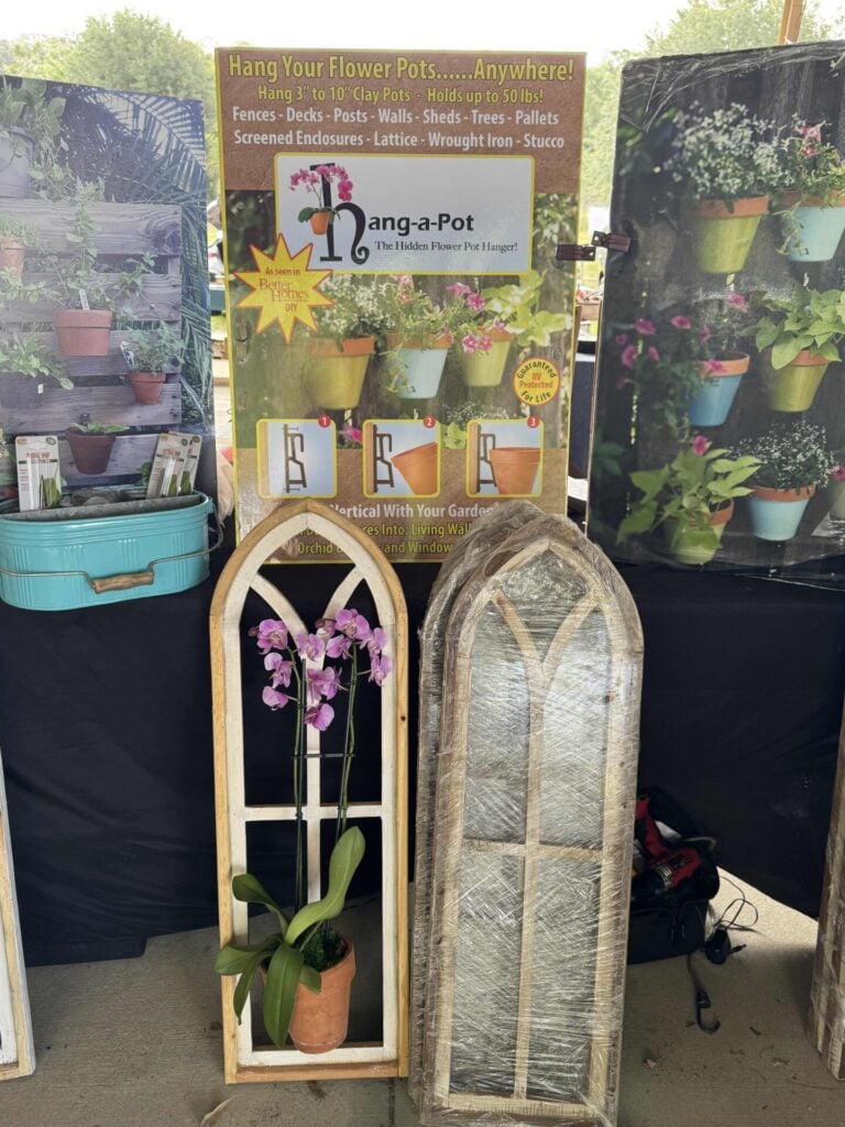 A display with a Hanga-Pot poster showing hanging flower pots, a turquoise container, and two gothic-style wooden frames—one with a potted orchid, the other wrapped in plastic.