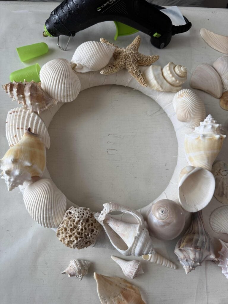 A wrapped wreath form featuring assorted seashells and a starfish sits on a table beside a hot glue gun and loose shells, showcasing a creative project in progress.