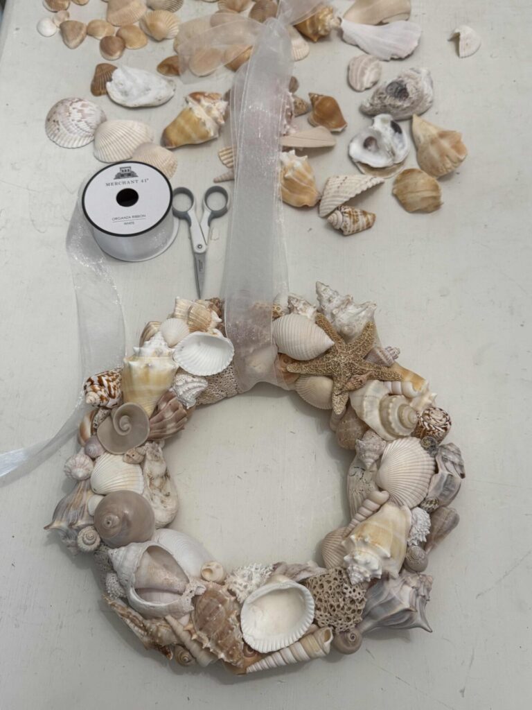 A DIY Shell Wreath made of assorted seashells and a starfish sits on a table, surrounded by loose shells, scissors, and a spool of white ribbon. Neutral-toned shells and a sheer ribbon add coastal charm to this handcrafted decor piece.