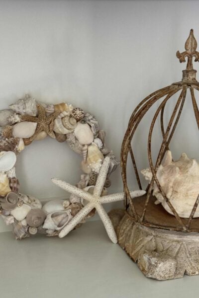 A handcrafted DIY Shell Wreath made of assorted seashells and starfish sits next to a conch shell inside a rustic, open metal cage on a stone base, all beautifully arranged on a light-colored surface.
