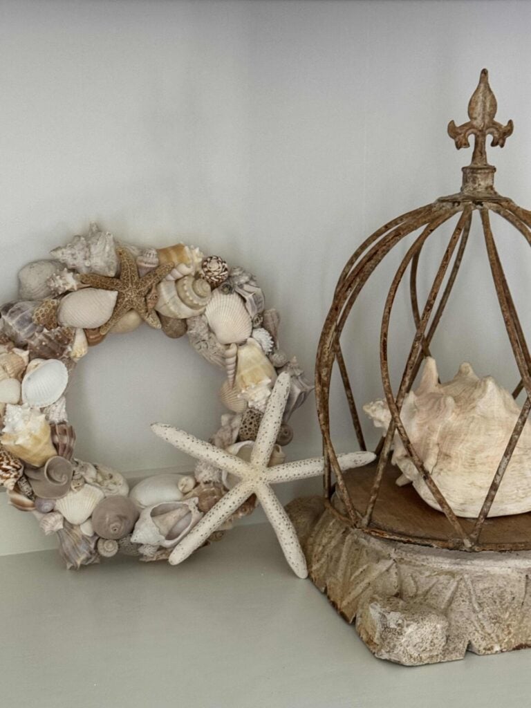 A handcrafted DIY Shell Wreath made of assorted seashells and starfish sits next to a conch shell inside a rustic, open metal cage on a stone base, all beautifully arranged on a light-colored surface.