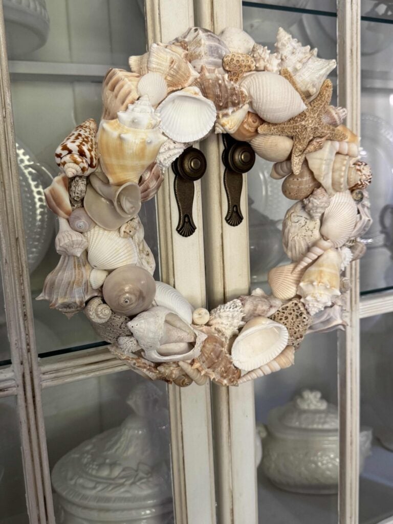 A decorative DIY Shell Wreath made of assorted seashells and a starfish hangs on the handles of a white cabinet with glass doors, displaying white ceramic dishes inside.
