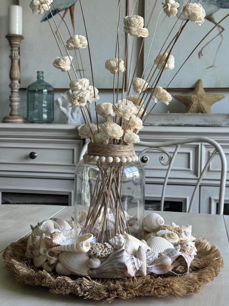 A glass jar filled with dried white flowers on long stems sits on a table, surrounded by seashells, coral, and a DIY Shell Wreath atop a round woven mat, creating a coastal-themed centerpiece in a light, airy room.