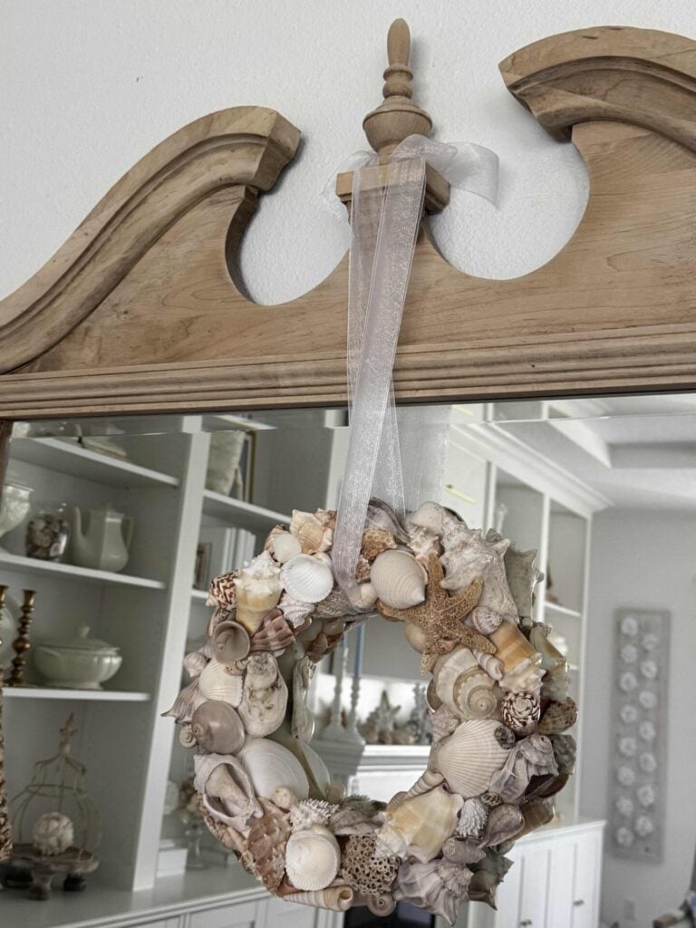 A DIY Shell Wreath made of assorted seashells hangs from a wooden mirror frame with a sheer white ribbon. In the background, white shelves display dishes and decor in a bright, airy room.