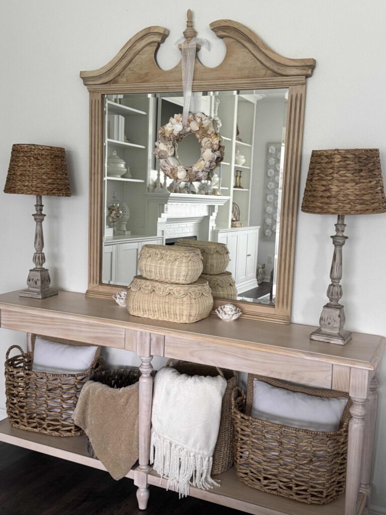 A wooden console table with woven baskets, blankets, and decorative pillows sits below a large mirror, offering charming inspiration for Summer Decor Ideas. Two wicker lamps and stacked woven baskets decorate the tabletop, while a seashell wreath hangs on the mirror to complete this breezy seasonal look.