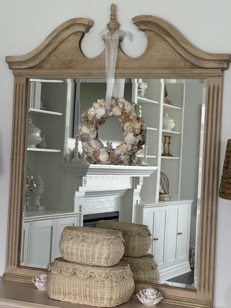 A decorative mirror with a wooden frame reflects a white room. A seashell wreath hangs on the mirror, and three stacked wicker baskets sit on the tabletop below, flanked by small shell decorations.