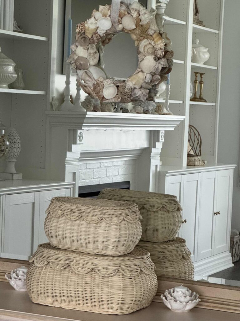 Three woven baskets are stacked in front of a white mantel and shelves. A decorative wreath made of seashells hangs above the mantel, and other white and neutral decor items are displayed on the shelves.